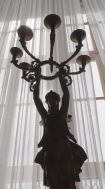 Mum heykelciğine yakın çekim. Başla. Vitrinde mum tutacağı olan lüks heykelcik. Pencereli lüks iç mekanda Avrupa tarzı heykel.