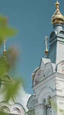 Ortodoks kilisesinin resimli görüntüsü. Şarjör. Beyaz kilisenin arka planında kubbeli yeşil dalların yakın çekimi. Yazın mavi gökyüzünün arka planında altın kubbeleri olan güzel beyaz kilise..