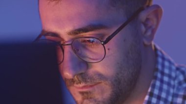 Bilgisayarın önünde çalışan yorgun genç adamın yakın çekimi. Medya. Gözlüklü genç adam bilgisayarda program yapıyor. Yorgun programcı bilgisayarda çalışıyor. 