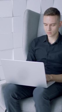 Modern minimalist koltukta oturan genç adam beyaz duvara karşı dizüstü bilgisayarla çalışıyor. Başla. Ofiste çalışan siyah gömlekli, yan manzaralı.