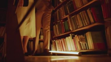 Karanlık bir kütüphanede yavaşça yürüyen bir çiftin alçak açılı görüntüsü. Stok görüntüleri. Kadın ve erkek kitaplarla rafların önünden geçiyor.