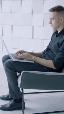 Modern minimalist koltukta oturan genç adam beyaz duvara karşı dizüstü bilgisayarla çalışıyor. Başla. Ofiste çalışan siyah gömlekli, yan manzaralı.