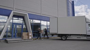 Lojistik perakende deposunun dışına bak, mal yükleme işlemi. Medya. İşçiler büyük karton kutuları teslimat kamyonuna koyuyorlar. 