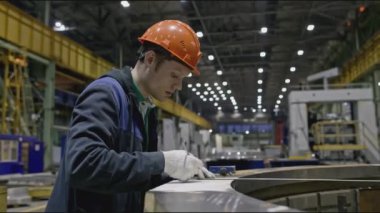 İşçi fabrikadaki yapısal parçaları ölçer. Borsa klipsi. Fabrikadaki miğferli işçiler parçaların ölçümlerini yapar. Genç adam fabrikada çalışıyor ve tasarım taş yapısını ölçüyor.. 