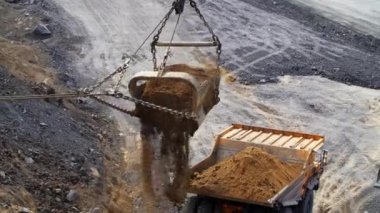 Aktif bir maden bölgesinde, bir kazıcı özenle toprağı hareket ettiriyor ve bekleyen bir çöp kamyonuna aktarıyor.