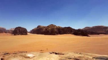 Kurak bir çöl manzarasının nefes kesici manzarası berrak mavi gökyüzünün altında kayalık dağları görkemli bir şekilde sergiliyor. Eylem