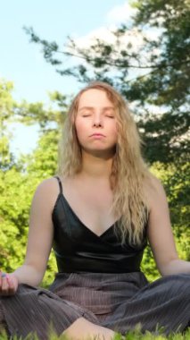 Genç, güzel, gevşemiş sarışın kadın yeşil çimlerde oturuyor ve meditasyon yapıyor. Kavram. Doğayla birleşme, şehir parkında meditasyon.