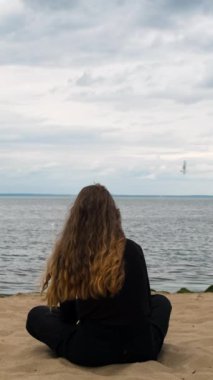 Finlandiya Körfezi, Saint Petersburg, Rusya 'daki bir kadının arka görüntüsü. Kavram. Siyah elbiseli, uzun saçlı, kumlu bir sahilde oturan ve uzaklara bakan genç bir kızın arka görüntüsü..