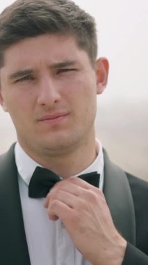Takım elbiseli genç adam dışarıda papyonuna dokunuyor. Başla. Smokin giyen adam sisli bir şekilde sahada dikiliyor.