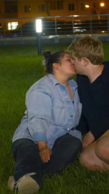 Çimlerde öpüşen genç bir çift. Medya. Geceleri parkta yeşil çimlerde oturan aşık bir çift öpüşüyor. Öpüşen mutlu şişman insanlarla dolu bir gece parkı..