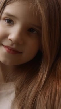 İçeride poz veren sevimli küçük bir kızın portresi. Stok görüntüleri. Güzel, gülümseyen kız uzun saçlarına dokunuyor ve pencerede poz veriyor.