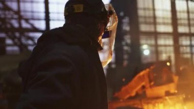 Güvenlik teçhizatı içinde çalışkan bir işçi loş bir endüstriyel ortamda metal eritme sürecini izliyor. Menkul Kıymetler