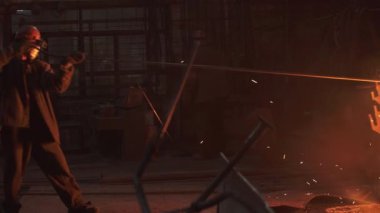 Koruyucu giyimli bir işçi, sönük bir fabrikada kıvılcımlar saçan uzun metal bir çubuğu ustalıkla tutuyor. Menkul Kıymetler