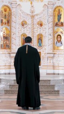 Iconostasis bir ortodoks kilisesinin içinde. Video kaydı. Azizlerin yüzlerinin, din kavramının, kilisenin içindeki iç ayrıntıların olduğu ikonların önünde duran bir rahibin arka görünüşü.