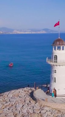 Deniz ve tatil köyünün arka planındaki deniz fenerinin en üst görüntüsü. Şarjör. Güzel beyaz deniz feneri mavi denizde duruyor ufukta şehri olan dağlara bakıyor..