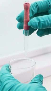 Şeffaf sıvının beyaz bir arka planda pipetle şişeye döküldüğü yakın plan. Şarjör. Eldivenli eller pipete bastırıyor.