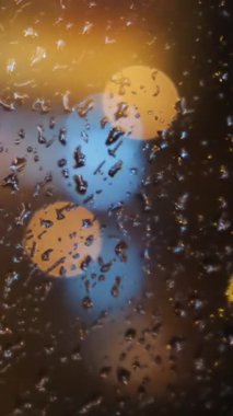 Penceredeki bulanık ışık noktalarının yakın çekimleri ve akşamları bardağa düşen yağmur damlaları. Kavram. Yağmurlu hava