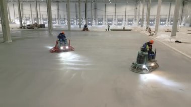 İki yetenekli işçi bir deponun içindeki beton zemini düzleştirmek ve düzleştirmek için makineler kullanıyorlar. Kırp