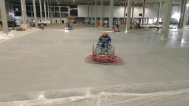 Kendini işine adamış bir işçi, büyük bir inşaat alanında taze beton döşemeyi pürüzsüzleştirmek için ustalıkla bir güç malası kullanır. Kırp