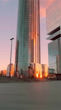 Camdan yapılmış modern bir iş merkezinin önündeki küçük bir meydan. Pencerelere yansıyan altın bir günbatımı. Video kaydı. Modern mimari kavramı