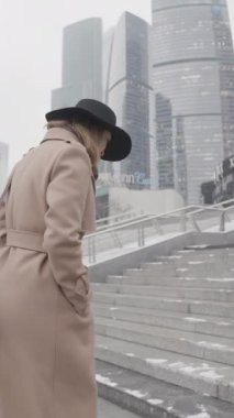 Kahverengi ceketli ve siyah şapkalı güzel iş kadını modern şehir bölgesinde gökdelenleri olan üst kata çıkıyor. Başla. Güzel cam cephe binalarının yanında yürüyen bir kadının yan görüntüsü.