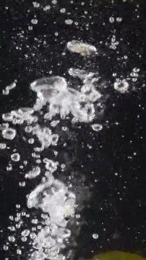 Limonun suya düşüşünü yakından çek. Başla. Tüm limon siyah arka planda kabarcıklarla suya düşer. Limon damlalarını suya dönüştürüyor..
