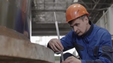 Sıkı bir şapka takan kendini işine adamış ve odaklanmış bir işçi, endüstriyel ortamdaki önemli ekipmanları titizlikle ölçüyor. Menkul Kıymetler