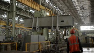 Bir sanayi işçisi geniş bir fabrikada ağır makineleri güvenli bir şekilde kaldıran büyük bir vinci aktif olarak denetliyor. Menkul Kıymetler