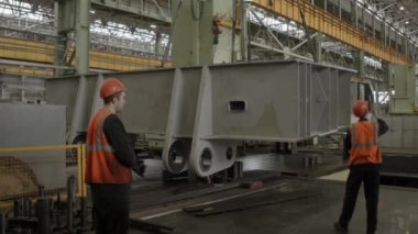 İşçiler koruyucu güvenlik teçhizatlarını ustalıkla giyip endüstriyel bir çalışma ortamında büyük metal bir nesneyi manevra ettiriyorlar. Menkul Kıymetler