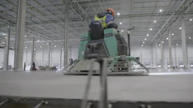 Bir işçi, geniş bir endüstriyel depoda, pürüzsüz bir yüzeyi güvence altına almak için beton bitirici kullanıyor. Kırp