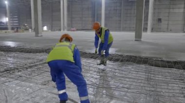 Geniş bir depo ortamında, kendini işine adamış iki işçi tesis için titizlikle beton bir zemin hazırlıyor. Kırp