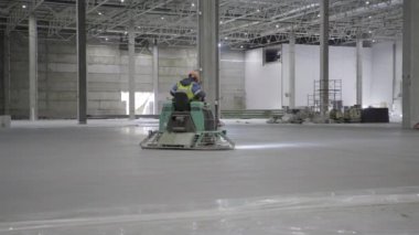 Kendini işine adamış bir işçi, geniş bir endüstriyel deponun içindeki zemin bitirme makinesini ustalıkla kullanıyor. Kırp