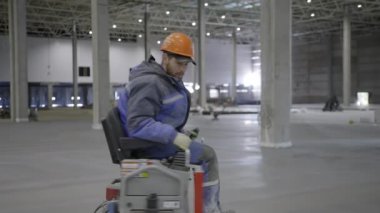 Çalışkan bir işçi büyük, ferah ve boş bir depo ortamında bir kat temizleyicisini ustalıkla çalıştırır. Kırp