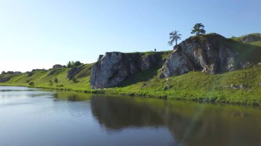 Güzel, sakin bir nehir sahnesinde açık mavi gökyüzünün altında kayalık oluşumların yanında yemyeşil tepeler sergilenir. Menkul Kıymetler
