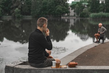 Huzurlu ve sakin bir göl manzarası. Bir adam bir şeyler atıştırırken, bir diğeri yakınlarda oturuyor.