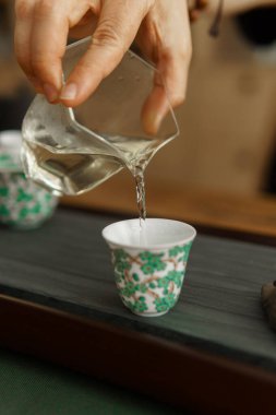Bir el zarif bir şekilde ahşap tepside duran zarif desenli bir çay bardağına temiz, aromatik bir sıvı döker.