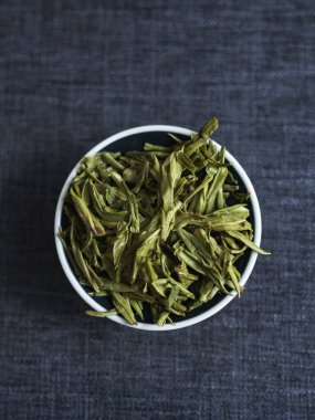 Kurumuş yeşil çay yapraklarının detaylı bir yakın plan görüntüsü sanatsal olarak seramik bir kasede koyu kumaş üzerine dizilmiş.