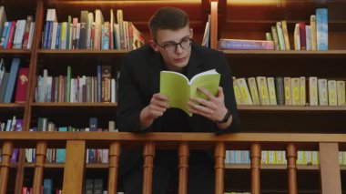 Kendini adamış ve odaklanmış genç bir adam, iyi stoklanmış bir kütüphane ortamında yeşil bir kitap okumaya dalmış durumda. Medya