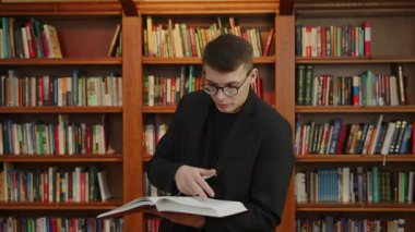 Genç bir adam renkli kütüphane raflarının canlı zeminine karşı yazılmış bir kitabı okumaya dalmış. Medya