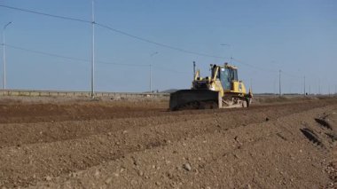 Gökyüzü açık ve aydınlıkken, büyük bir buldozer aktif bir şekilde tarıma hazırlanıyor. Kırp