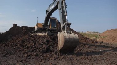 Bir kazıcı açık mavi gökyüzünün altındaki işlek bir inşaat sahasında toprak ve toprağı aktif olarak hareket ettiriyor. Kırp