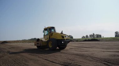 Parlak sarı bir yol silindiri bir inşaat sahasında çalışarak zemini asfaltlamaya etkin bir şekilde hazırlıyor. Kırp