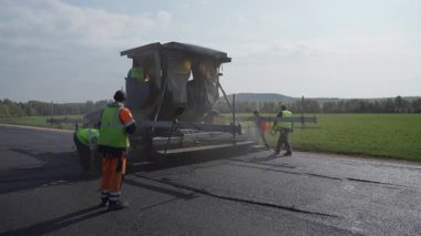 İşçiler, açık bir arazide açık mavi bir gökyüzünün altında asfalt döşeyecek ağır makineleri ustalıkla kullanırlar. Kırp
