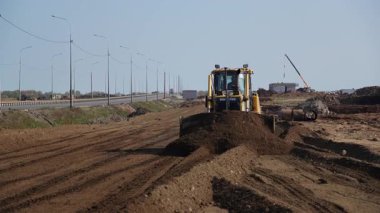 Bir inşaat sahasında, bir kazıcı toprağı kazıp hareket ettirirken, elektrik hatları arka plan boyunca uzanıyor. Kırp