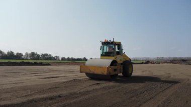 Güçlü bir silindir zemini yukarıdaki açık mavi gökyüzünün altındaki işlek bir inşaat sahasında sıkıştırıyor. Kırp
