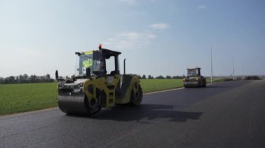 Dinamik bir sahne silindirleri etkin bir şekilde yepyeni bir yola zemin hazırlıyor. Etrafı canlı yeşil alanlarla çevrili. Kırp