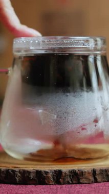 Kırsal bir ahşap masadaki cam çaydanlığa buharlı çay döken bir insanın sakin sahnesi, ritüelin güzelliğini ve berraklığını vurguluyor. Medya