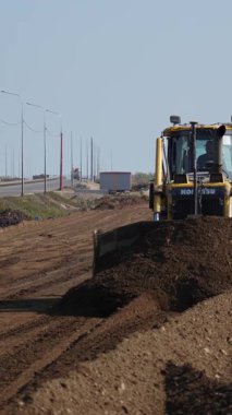 Bir inşaat sahasında, bir kazıcı toprağı kazıp hareket ettirirken, elektrik hatları arka plan boyunca uzanıyor. Kırp