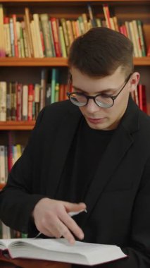 Genç bir adam renkli kütüphane raflarının canlı zeminine karşı yazılmış bir kitabı okumaya dalmış. Medya