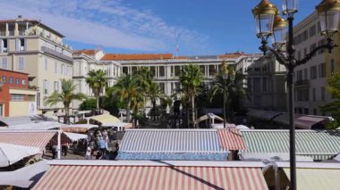 Cours Saleya pazarı, vizyonun tam ortasında, renkli tenteleri, palmiye ağaçları ve hareketli müşterileri olan canlı bir atmosfer sunar.
